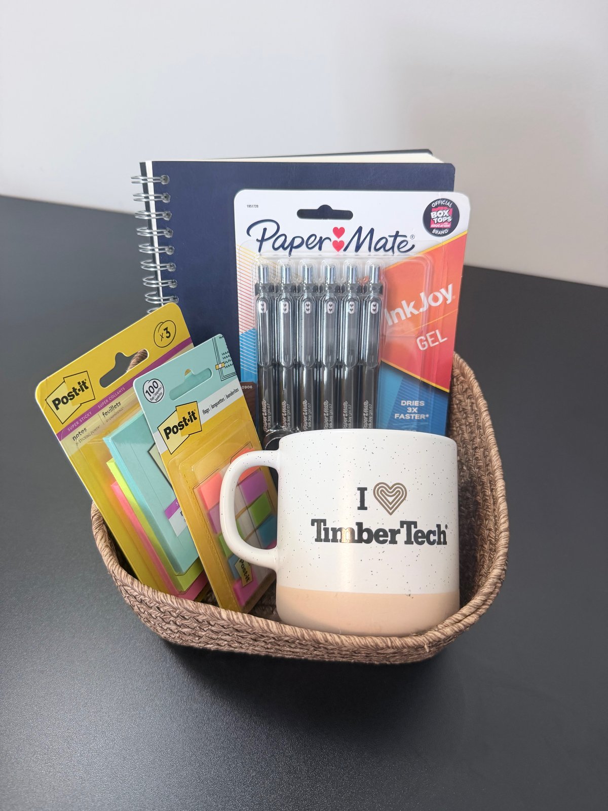 Corporate gift basket with a company-branded mug, notebook, gel pens, and desk supplies in a woven basket